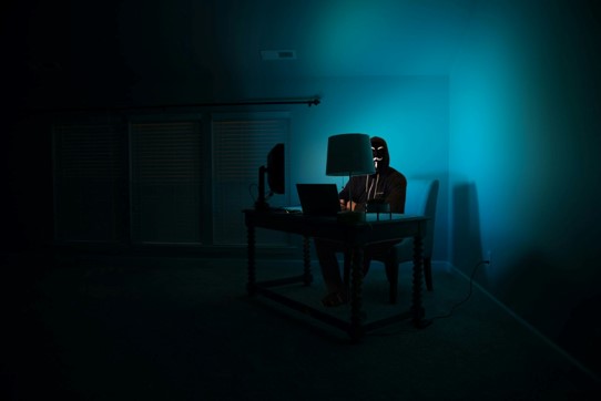 A silhouette of a person wearing a mask while working on a computer in a dark room, evoking ideas of online anonymity and data theft