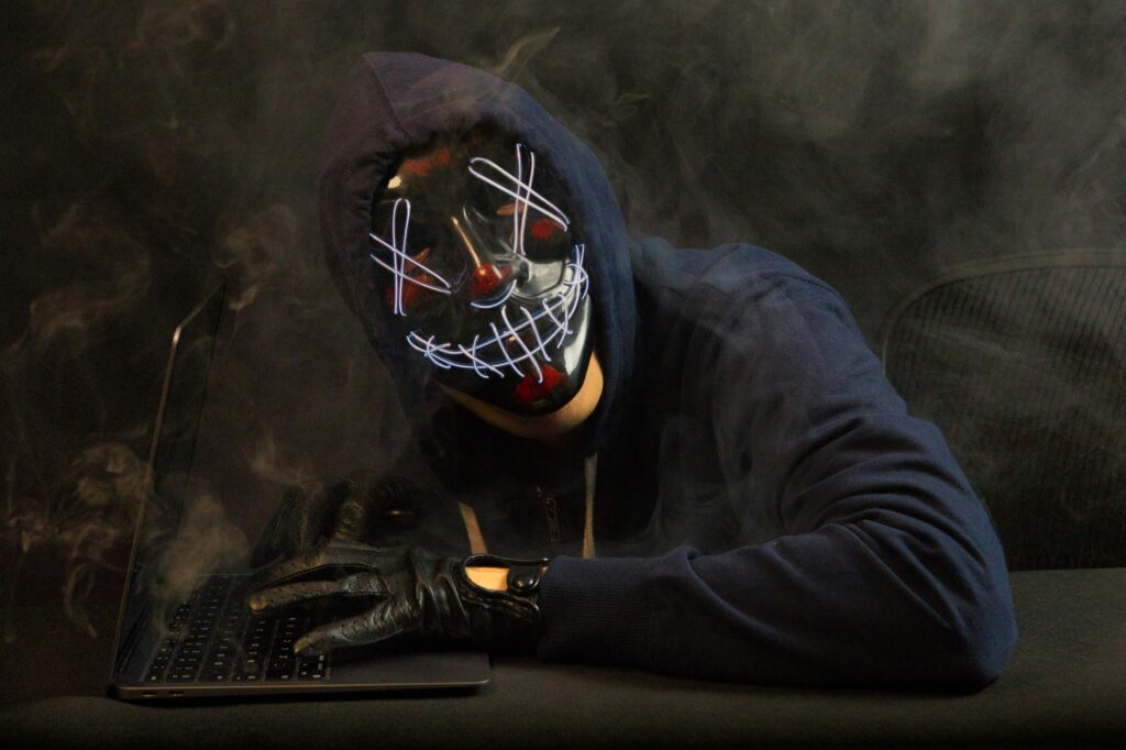 A person wearing an LED light-up mask with a sinister design, seated at a desk in a dark
