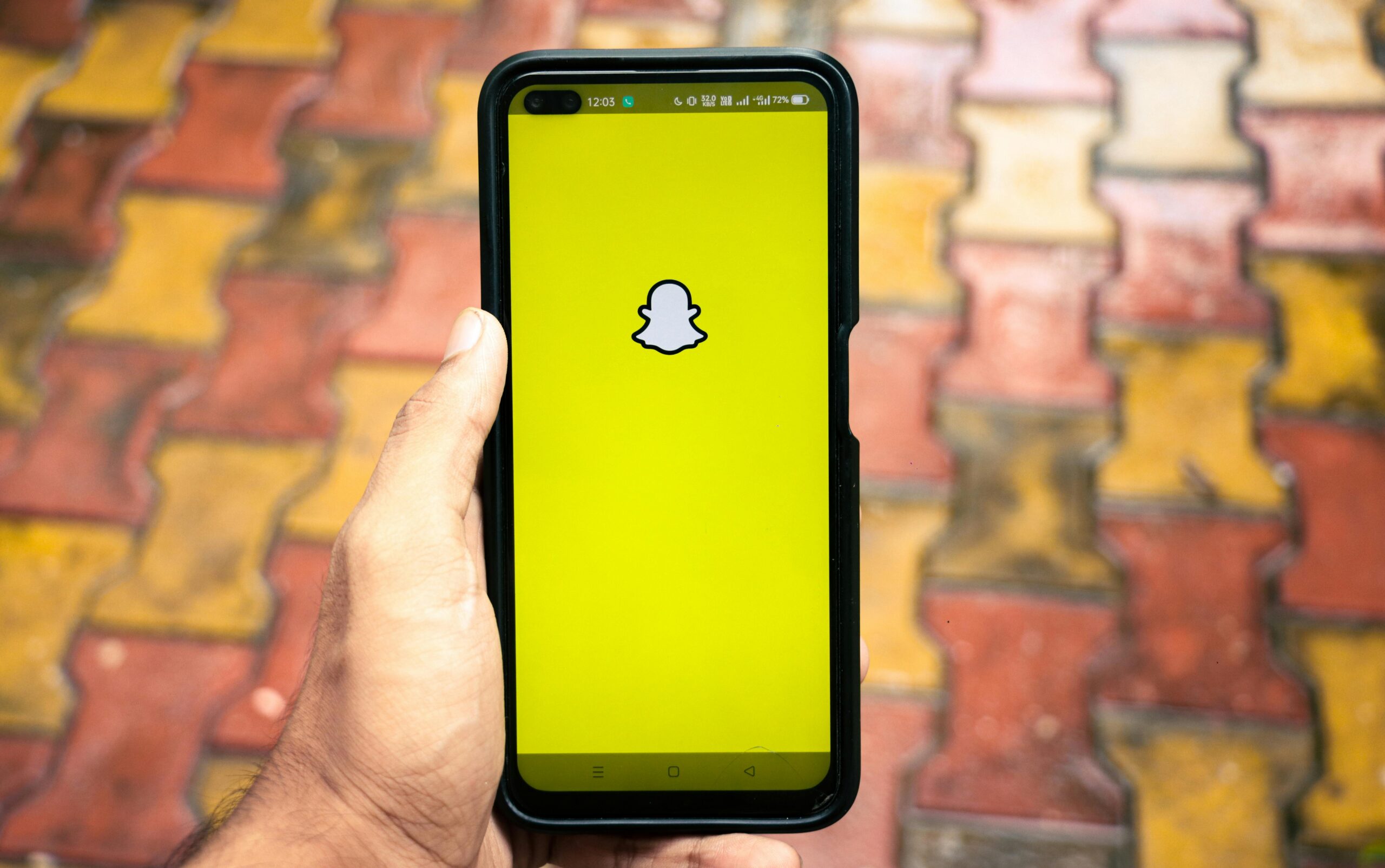 The image shows a person holding a smartphone with the Snapchat app open, displaying its characteristic yellow screen and white ghost logo