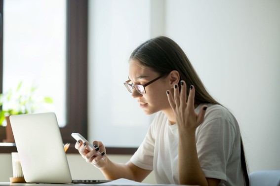 A young woman, wearing glasses, appears frustrated while looking at her smartphone, gesturing with her hand, with a laptop open in front of her on a desk