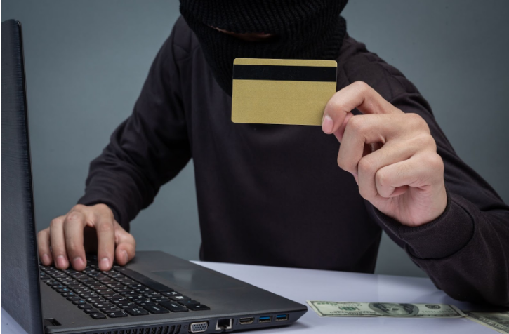 [Person wearing a mask holding a credit card while using a laptop, indicating credit card fraud or identity theft