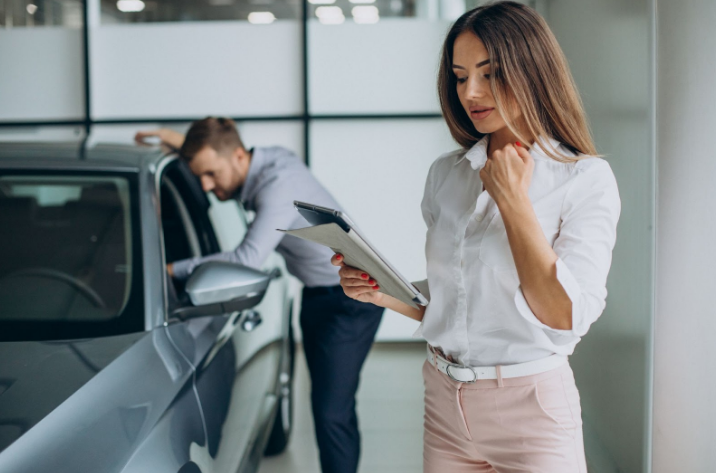 A woman with a clipboard reviews details while a man examines a car in a dealership showroom