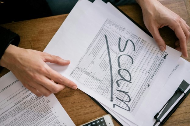 Close-up of hands holding documents on a clipboard with the word 'SCAM' written across a form, emphasizing the fraudulent nature of the paperwork