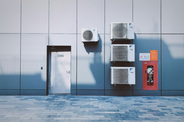 An outdoor setup with multiple wall-mounted HVAC units, a metal door, and a red fire hose cabinet against a sleek modern building exterior