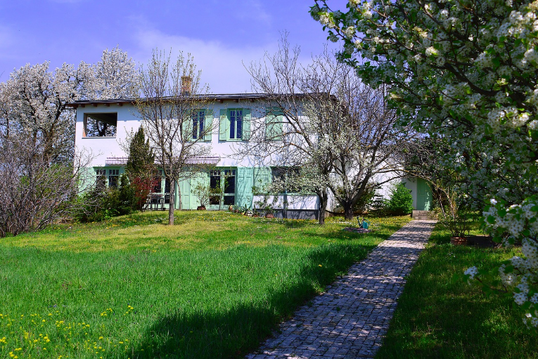 A white house with green shutters surrounded by a garden, blooming trees, and a stone pathway leading to the front entrance