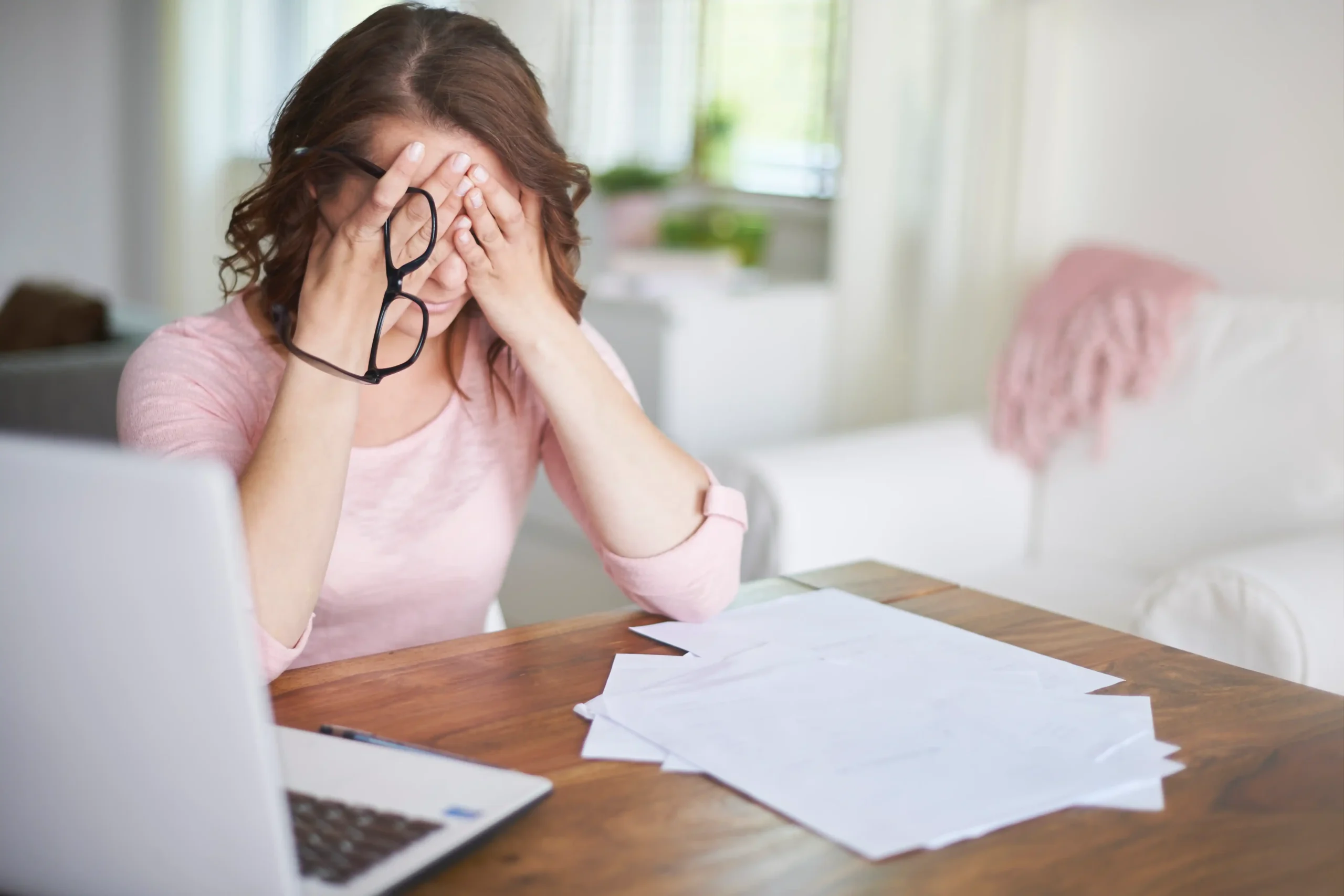 Stressed woman holding glasses, financial documents on table, worried about PPP fraudsters and business scams