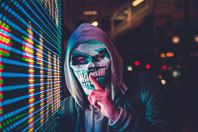 Person in hoodie and skull mask standing by a digital screen, symbolizing anonymous cyber threats