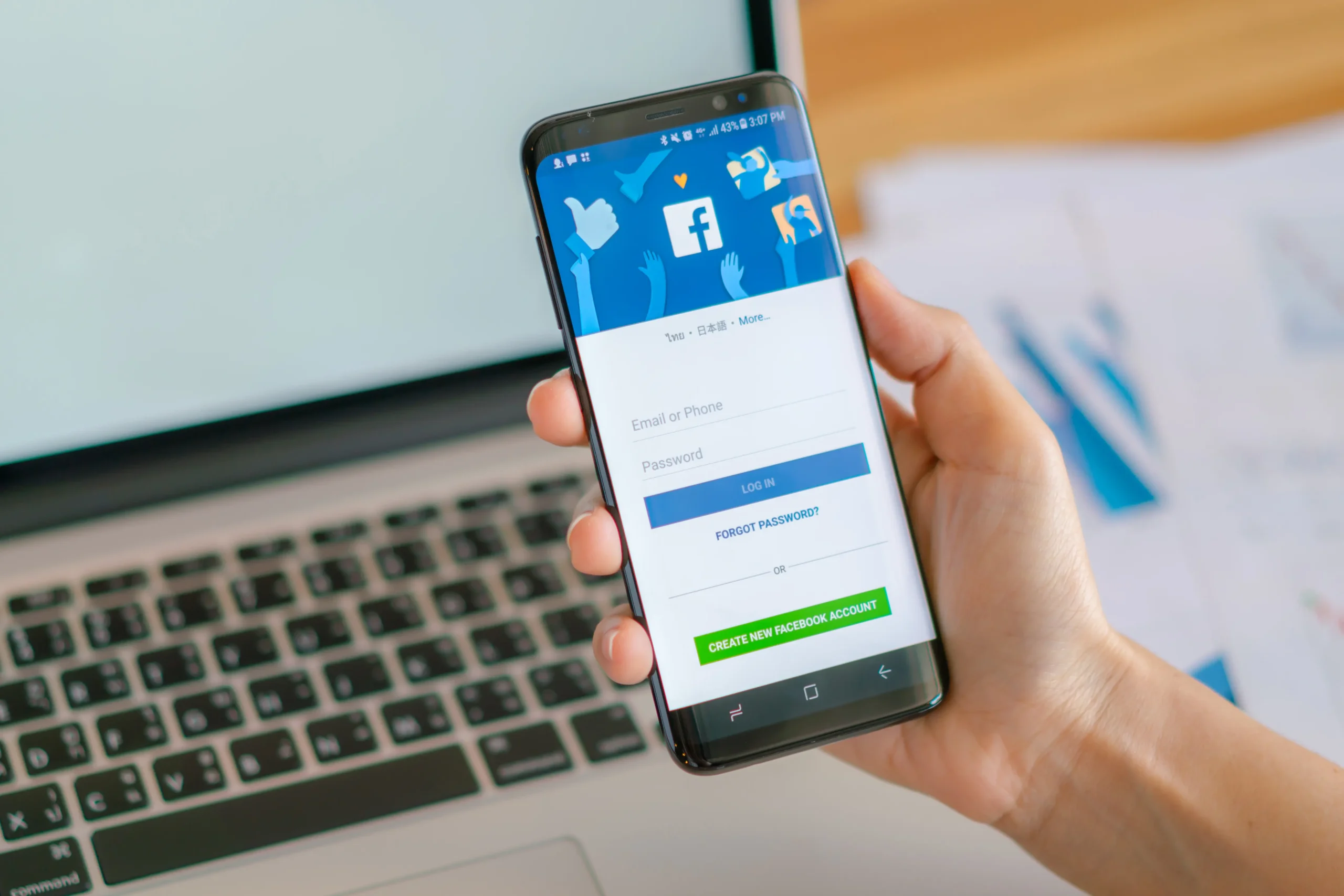 A person holding a smartphone with the Facebook login screen open, next to a laptop on a desk