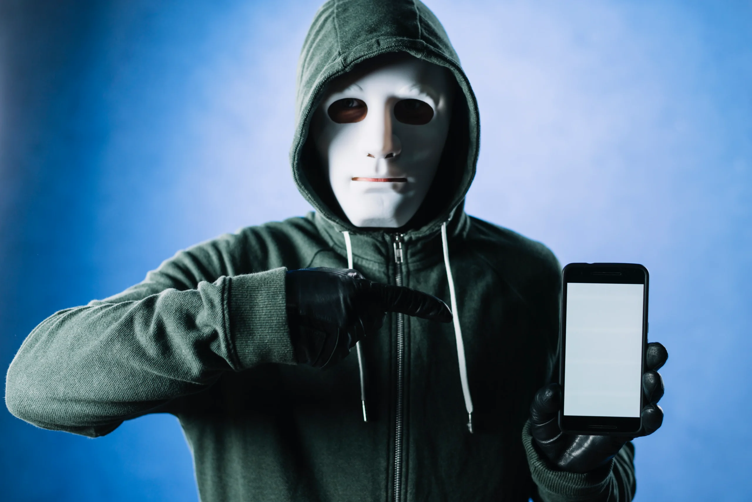 A scammer in a hoodie and white mask holding a smartphone